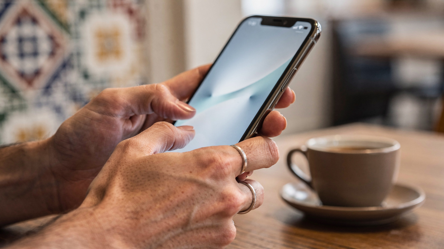 Close-up of a traveler's hands holding an iPhone 17, demonstrating the physical gesture of tapping through settings to activate an eSIM, with blurred cafe background.
