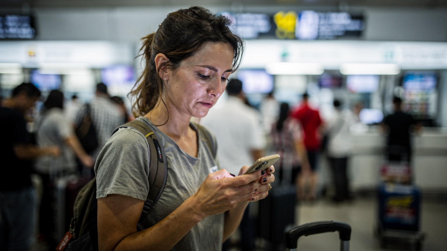 Bir seyahat blog yazarı, yerel bir SIM kart satın alırken uzun bir kuyrukta bekliyor, havaalanının yoğun ve biraz stresli atmosferi içinde telefonuna bakıyor.
