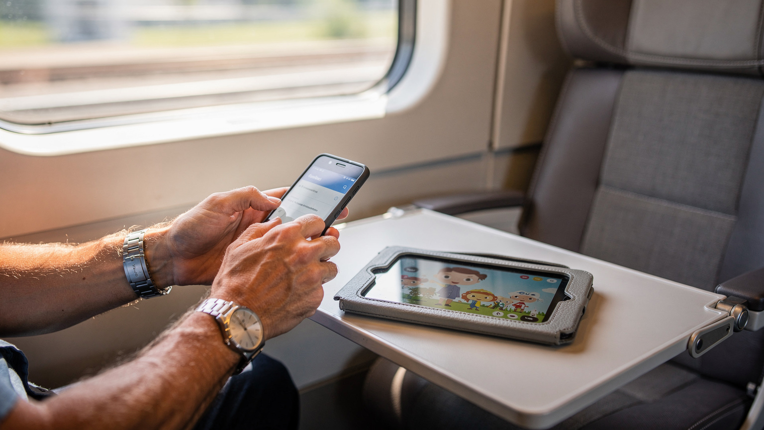 A parent's hands are configuring an older Android phone to act as a mobile hotspot, while a child's tablet, displaying a downloaded cartoon, rests on a tray table in the background, inside a clean, modern European high-speed train carriage.